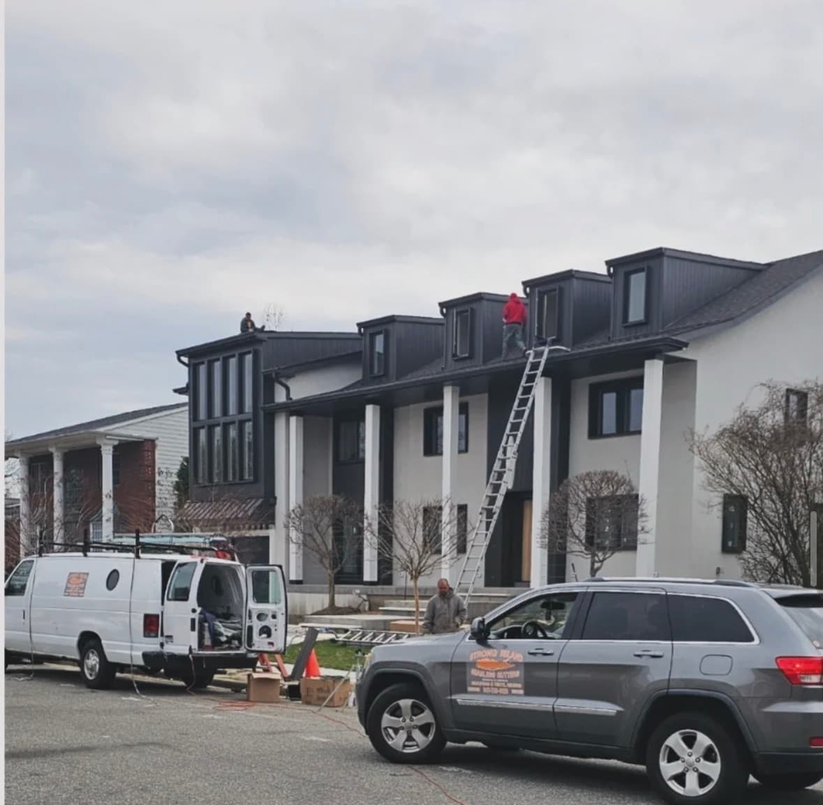 Two Strong Island Exteriors crew members working along the dormers of a dark-roofed home, with an extension ladder up to the roofline.